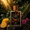 A bottle of Khus Ultimatum perfume by Ambassador Est. London stands in a dark, richly detailed natural setting with sunlight streaming through, alongside an orange, a halved orange, and pink flowers.